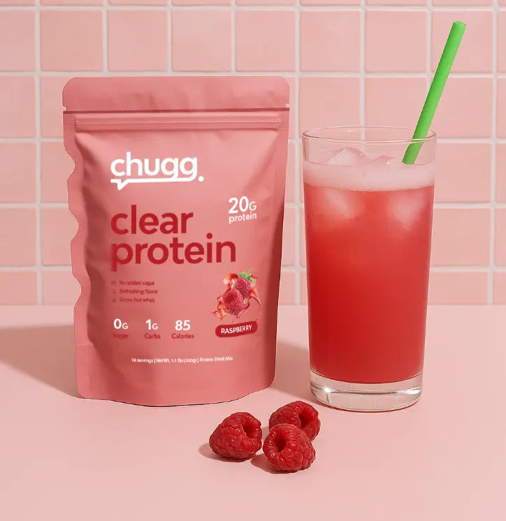 Chugg Clear Protein raspberry pouch next to a glass of raspberry protein drink with ice, green straw, and fresh raspberries on a pink surface