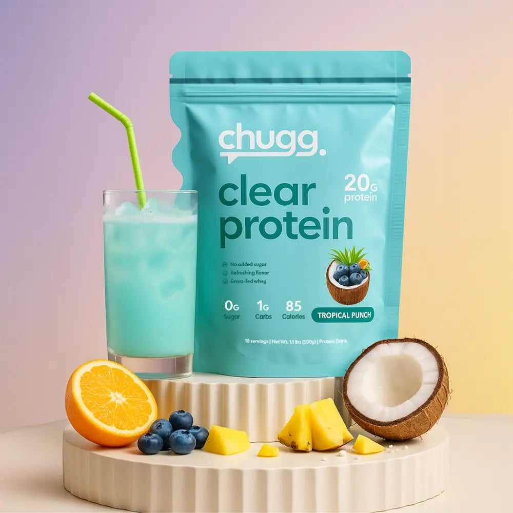 Chugg Clear Protein tropical punch pouch next to a glass of blue protein drink with green straw, surrounded by pineapple, orange, coconut, and blueberries on a pedestal