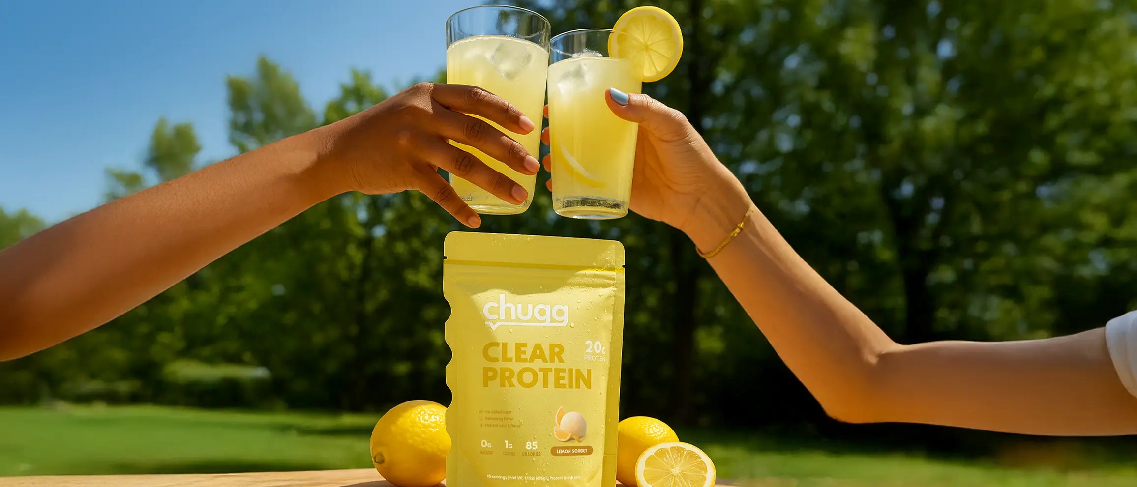 Two people clinking glasses of Chugg Protein Lemon Sorbet Flavored with a Clear Protein bag in the foreground, outdoors. Bright and refreshing